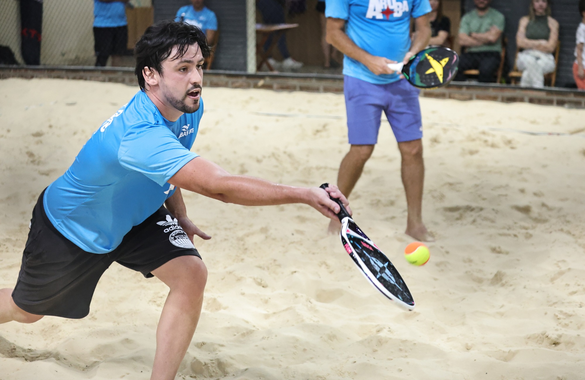 Atletas competindo no 1º torneio de Beach Tennis do Festuris em Gramado, com quadras de areia e público animado.