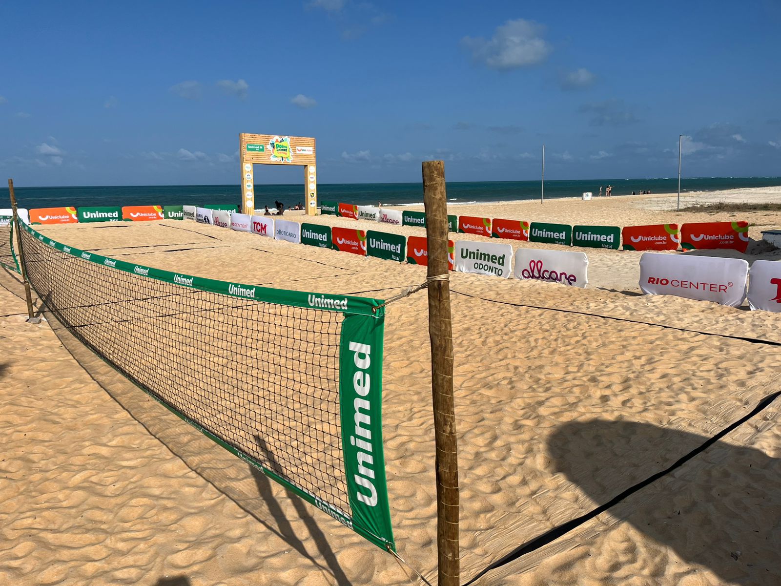 Participantes do Unimed Pirangi Open de Beach Tennis jogando em uma quadra de areia ensolarada.